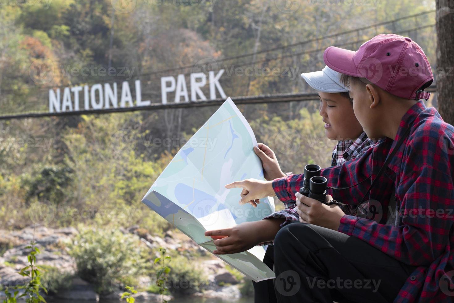 Asian young teen boys hold national park map, reading details of birdwatching before using their binoculars to watch the bird which sitting on brunches and flying in the sky, summer vacation concept. photo