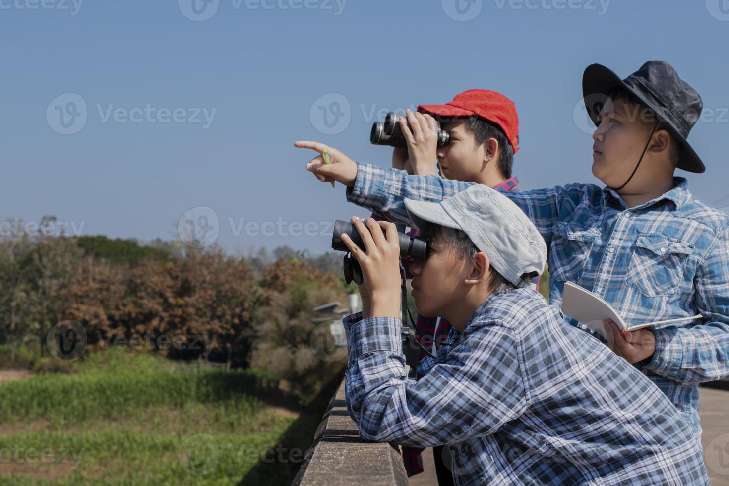 Asian boys using binoculars to watch birds on trees and fish in river in local national park during summer camp, idea for learning creatures and wildlife animals and insects outside the classroom. photo