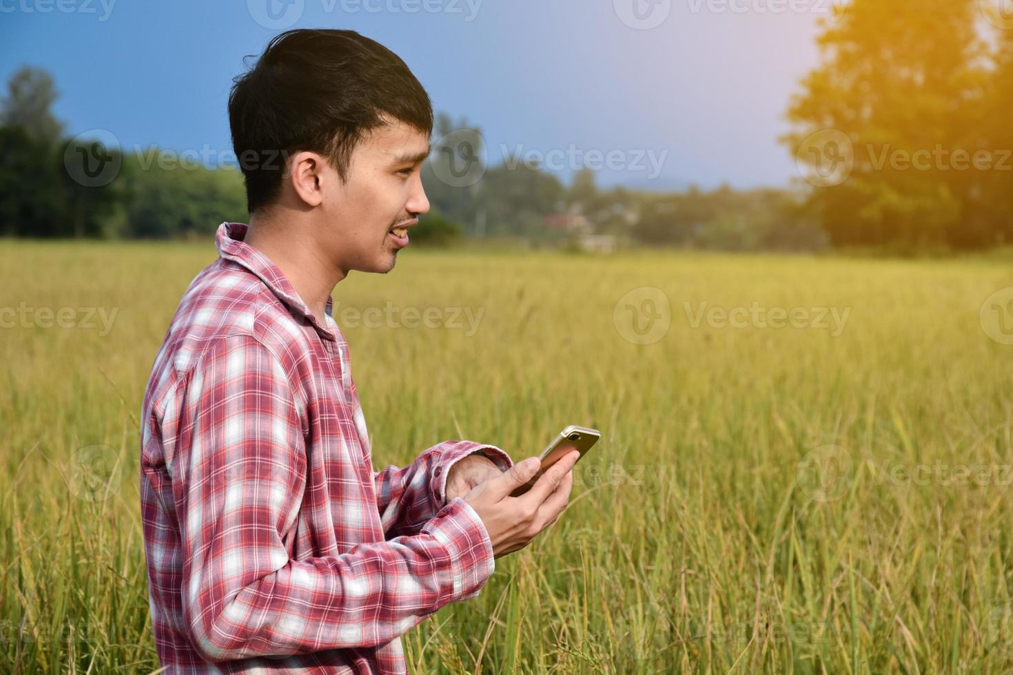 Portrait of young asian boy holds smart phone, standing in middle area of rice paddy farmland to give growing information, soft focus. photo