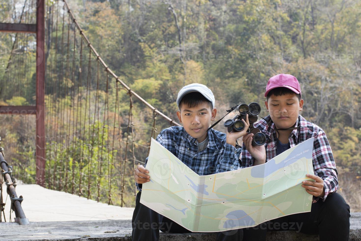 Asian young teen boys hold national park map, reading details of birdwatching before using their binoculars to watch the bird which sitting on brunches and flying in the sky, summer vacation concept. photo