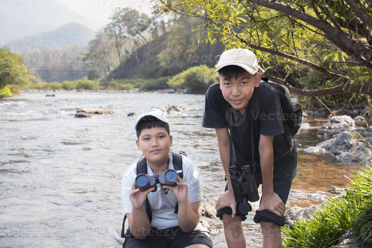 Asian boys using binoculars to watch birds on trees and fish in river in local national park during summer camp, idea for learning creatures and wildlife animals and insects outside the classroom. photo