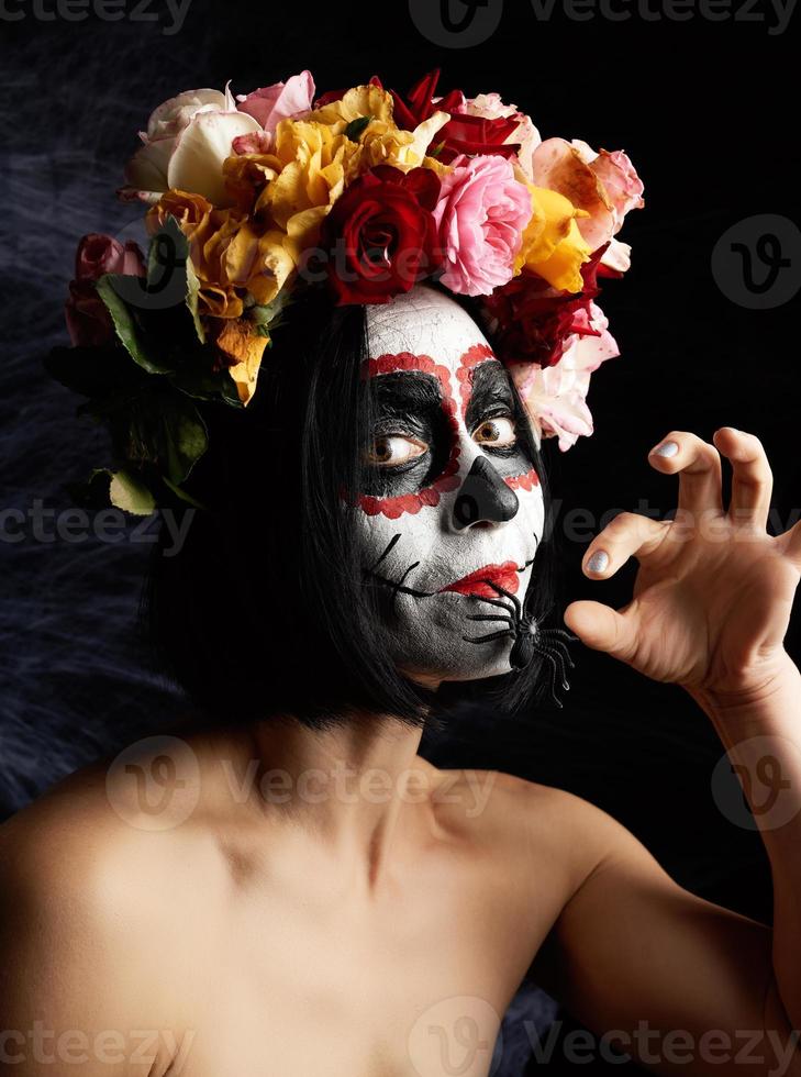 la chica con el pelo negro está vestida con una corona de rosas multicolores y el maquillaje está hecho en su cara calavera de azúcar hasta el día de los muertos foto