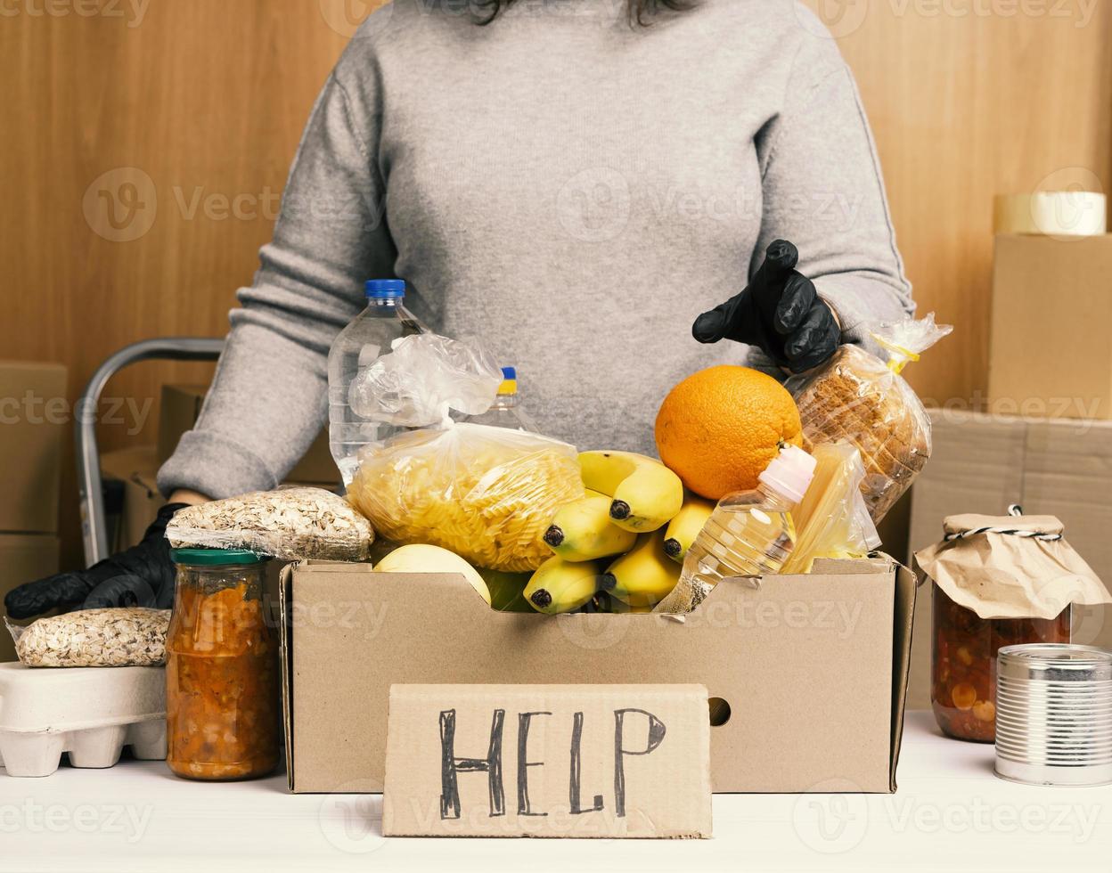 woman in gray sweater and black gloves holding a cardboard box with groceries, concept of assistance and volunteering photo