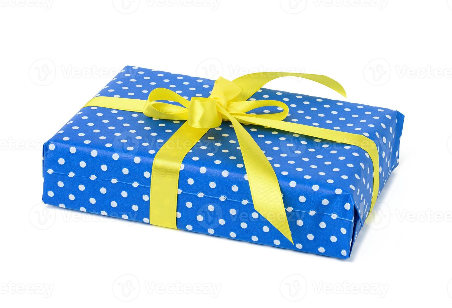 caja rectangular envuelta en papel azul con lunares blancos y atada con cinta de seda amarilla sobre fondo blanco foto