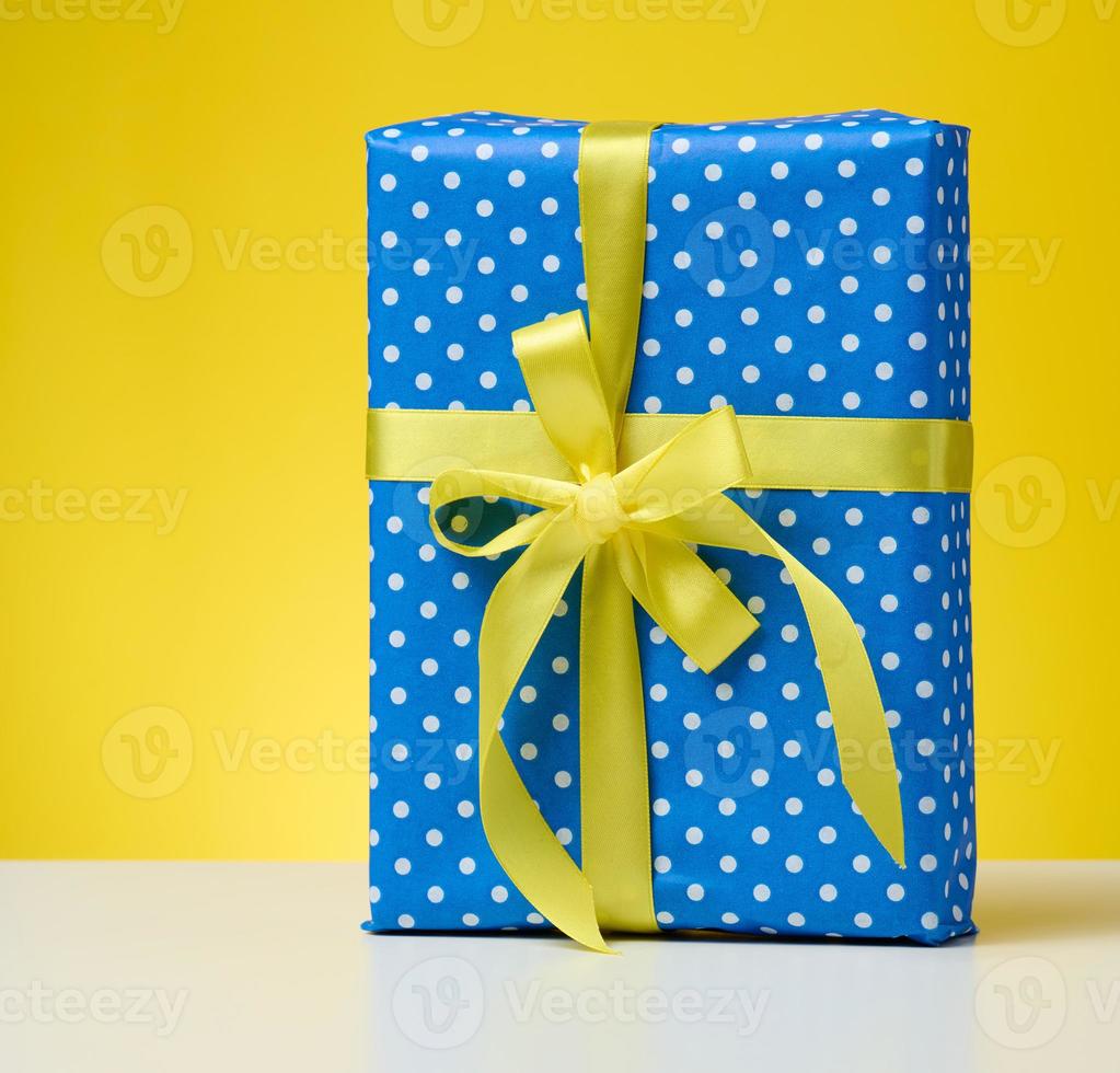 box packed in festive blue paper and tied with silk yellow ribbon on a yellow background, birthday gift, surprise photo