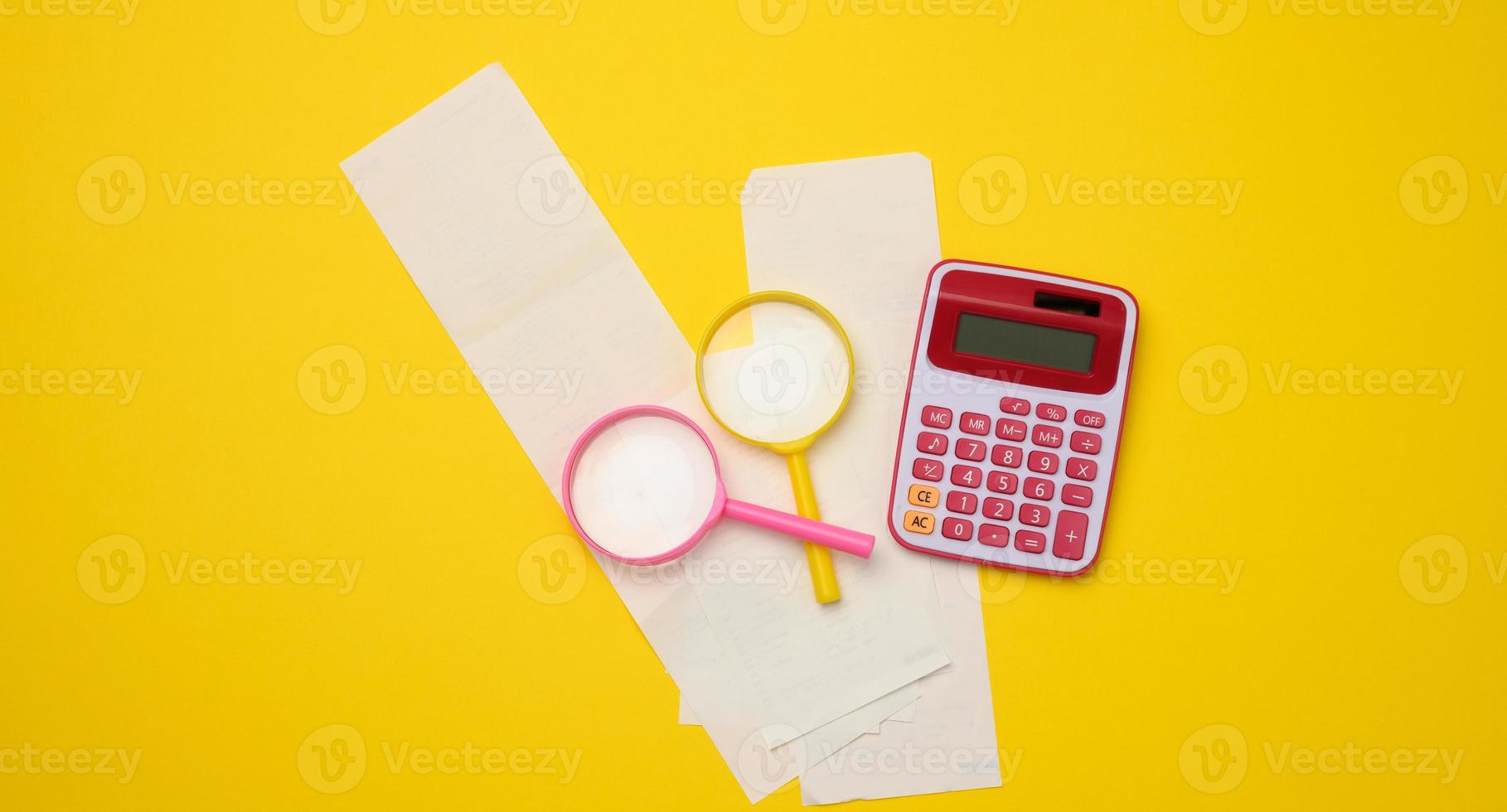 a stack of paper checks, a pink plastic calculator and a magnifying glass on a yellow background. Family budget audit concept photo
