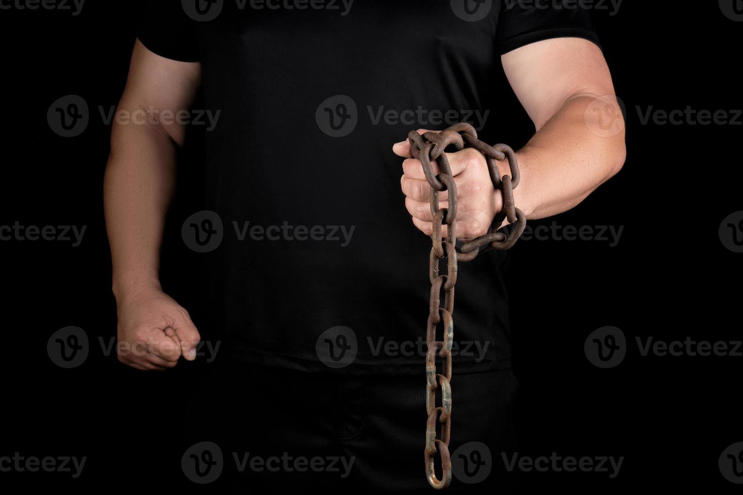 adult man in black clothes stands upright with strained muscles and holds a rusty metal chain photo