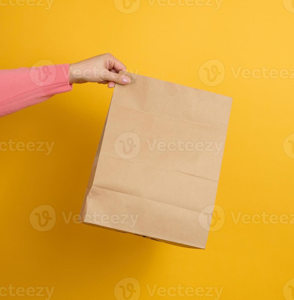woman holding in hand brown blank craft paper bag for takeaway on yellow background. Packaging template mock up. Delivery service concept photo