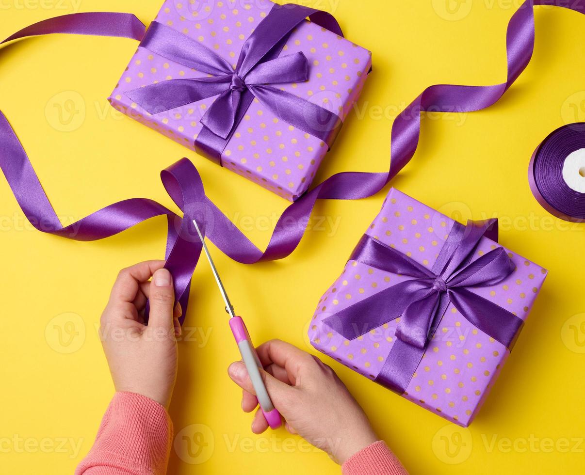 female hands are cutting a silk purple ribbon with scissors, next to gift boxes. Yellow background, top view photo