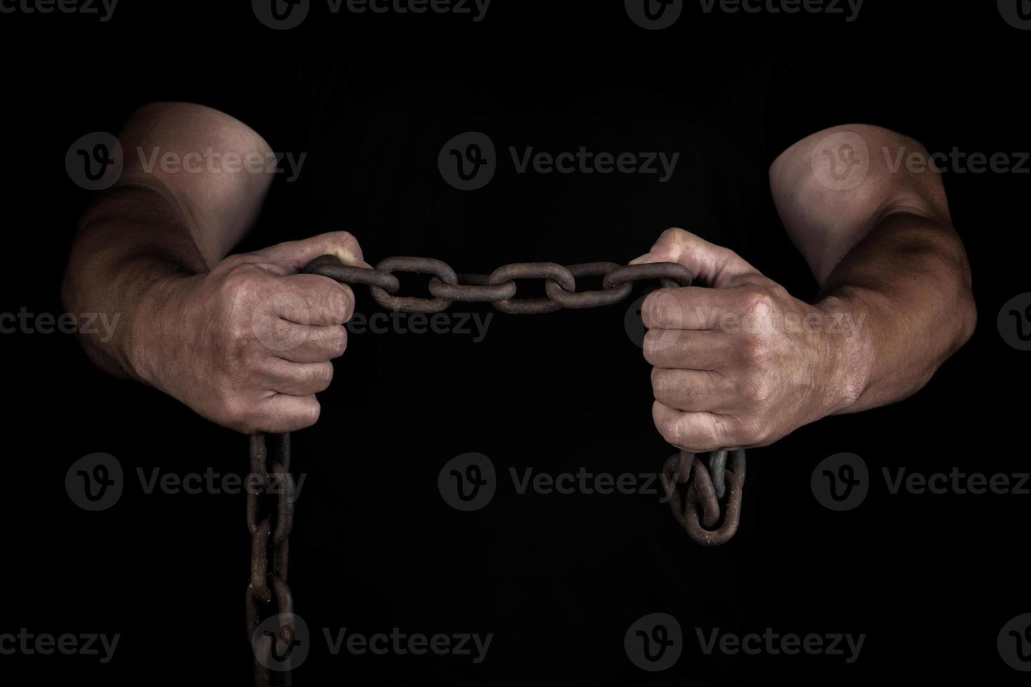 adult man in black clothes stands upright with strained muscles and holds a rusty metal chain photo