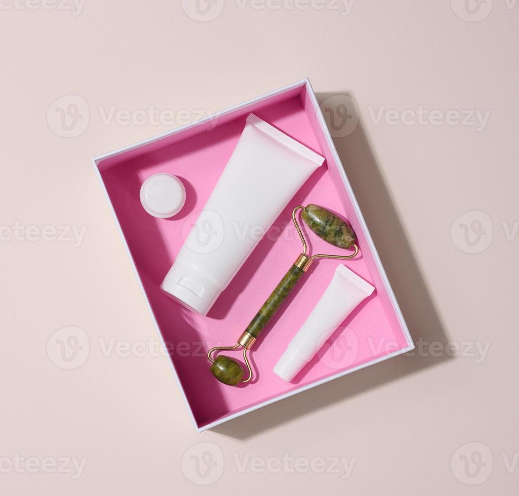 Jade manual facial massager and white plastic tubes for cosmetics on a beige background, top view photo