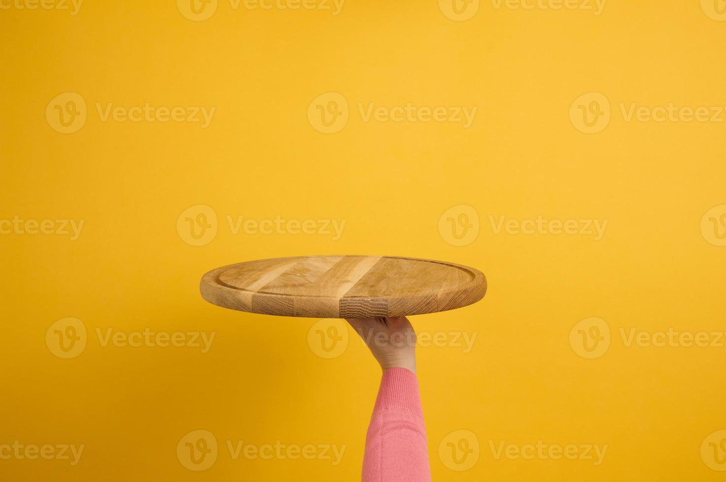 mujer sosteniendo una tabla de pizza de madera redonda vacía en la mano, parte del cuerpo sobre un fondo amarillo foto