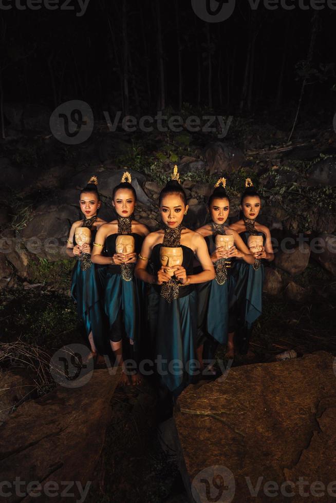 A Bunch of girls in green costumes holding a wooden masks in their hands while standing between the big rocks in front of the forest photo