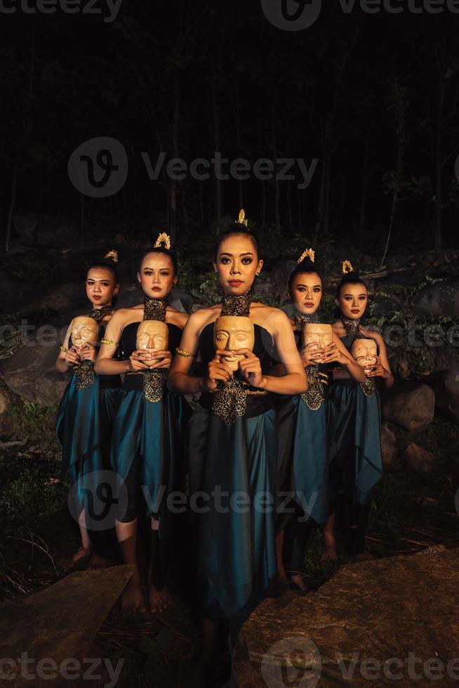 A Bunch of girls in green costumes holding a wooden masks in their hands while standing between the big rocks in front of the forest photo
