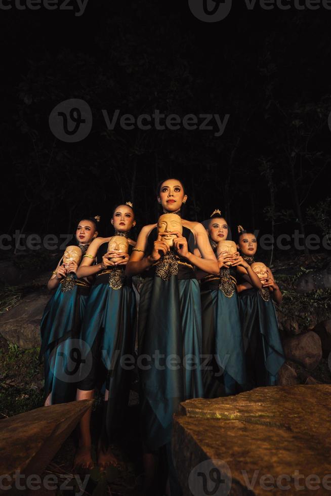 A Bunch of girls in green costumes holding a wooden masks in their hands while standing between the big rocks in front of the forest photo