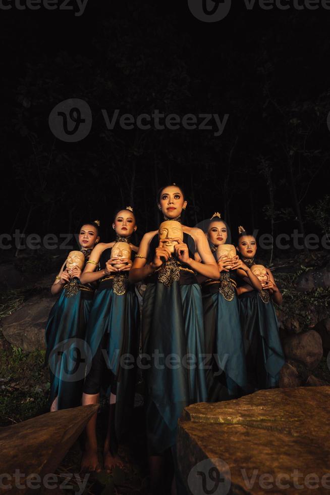 A Bunch of girls in green costumes holding a wooden masks in their hands while standing between the big rocks in front of the forest photo