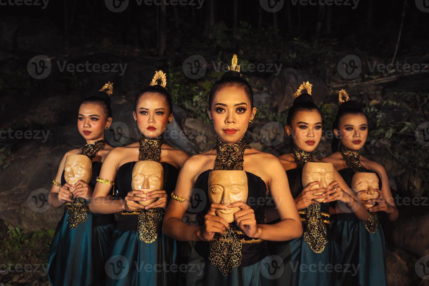 A Bunch of girls in green costumes holding a wooden masks in their hands while standing between the big rocks in front of the forest photo
