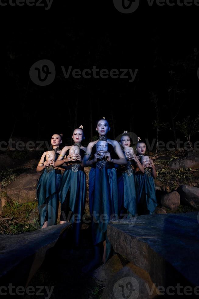 A Group of Javanese women standing in the woods between the rocks while holding a wooden mask photo