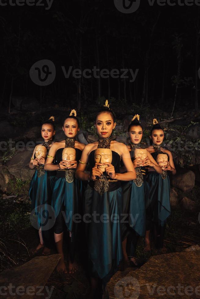 A Bunch of girls in green costumes holding a wooden masks in their hands while standing between the big rocks in front of the forest photo