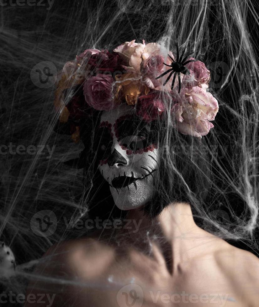 la chica con el pelo negro está vestida con una corona de rosas multicolores y el maquillaje está hecho en su cara calavera de azúcar hasta el día de los muertos foto