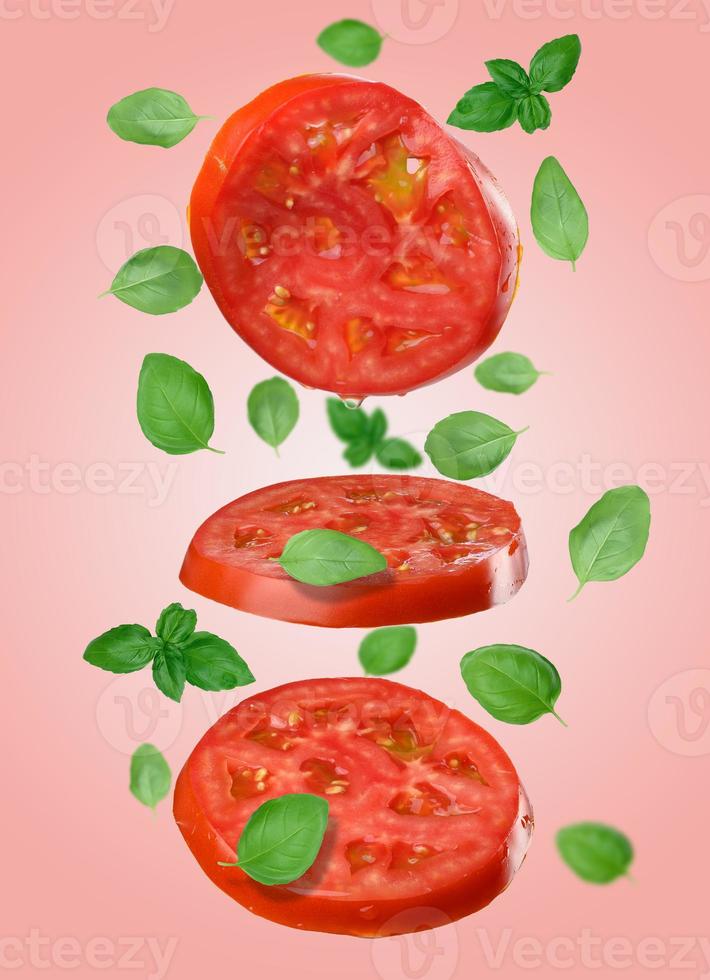 Round red slices of tomato and basil leaves levitate on a pink background. photo