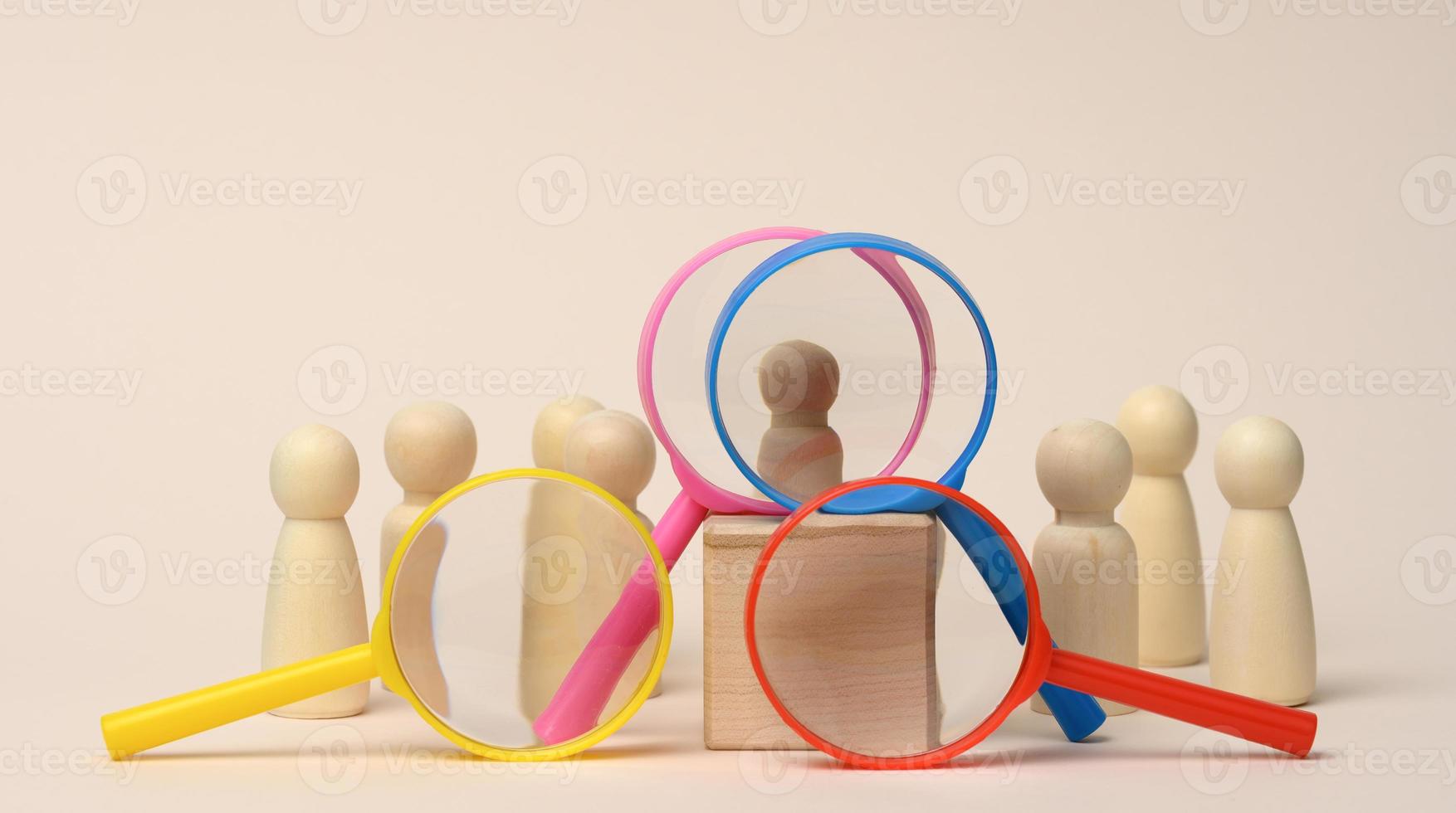wooden figures of men stand on a beige background and a plastic magnifying glass. Recruitment concept photo