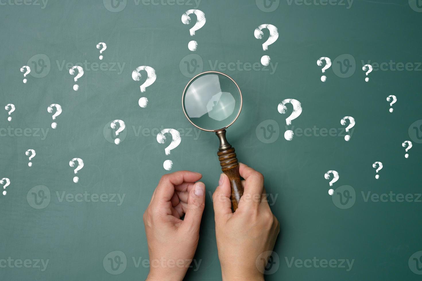 Female hand holds a plastic magnifying glass and question marks on a green background. photo