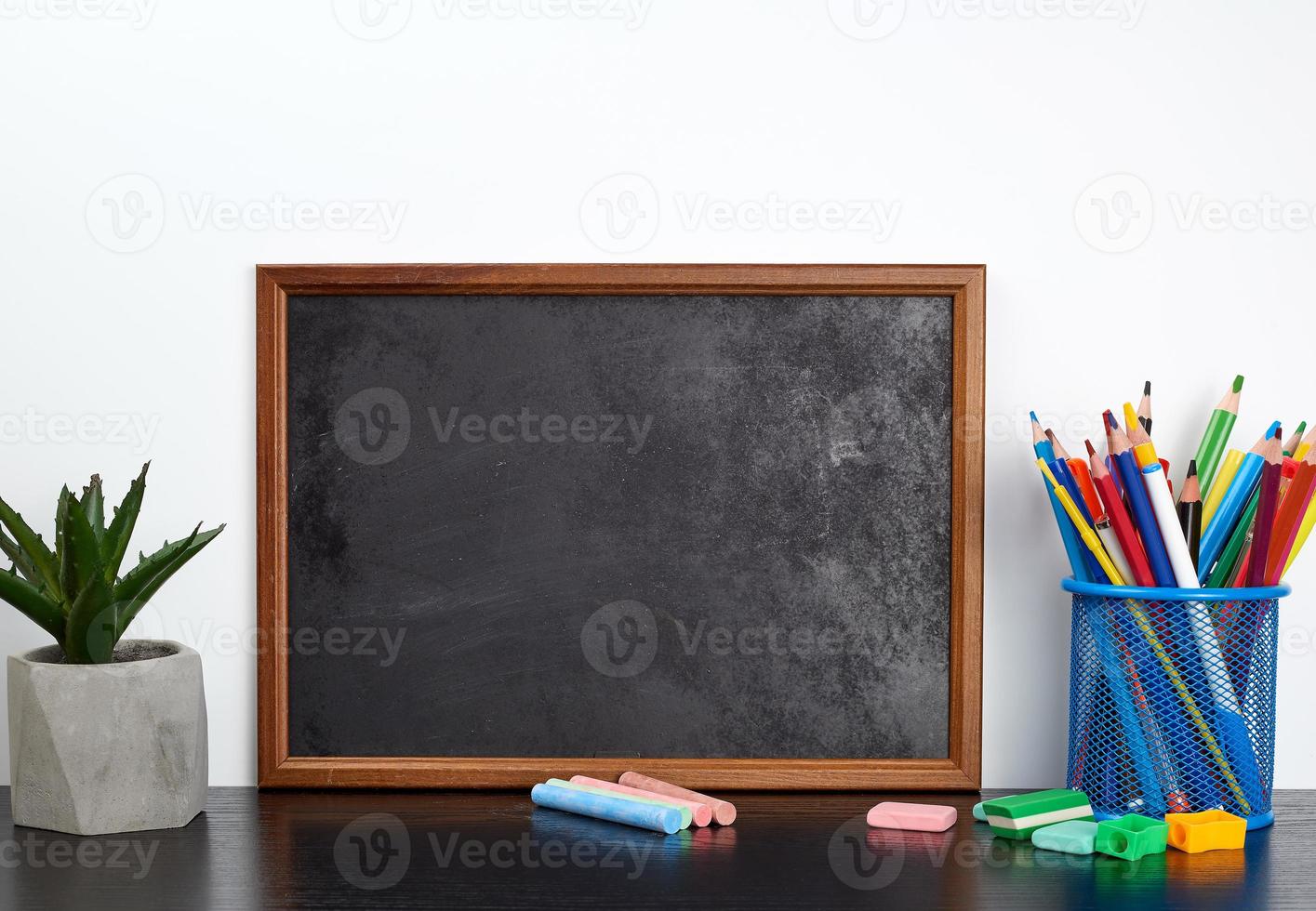 empty black chalk board, multi-colored pencils in a blue metal stand on a black table photo