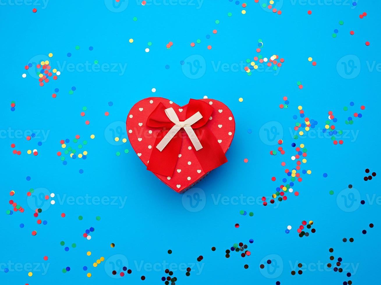 closed red heart-shaped cardboard box on a blue background with multi-colored shiny confetti photo