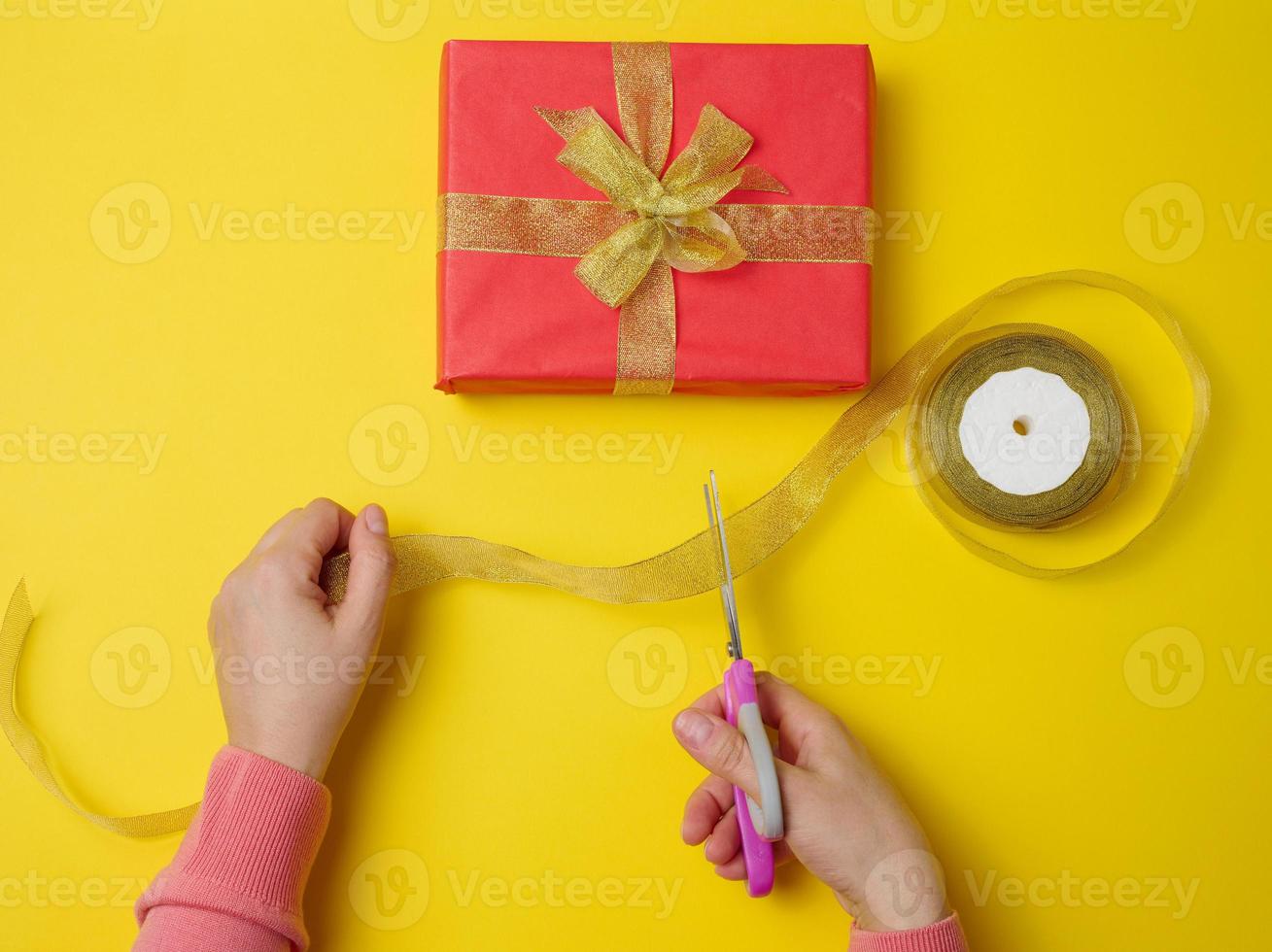 female hands are cutting a silk ribbon with scissors, next to gift boxes. Yellow background, top view photo