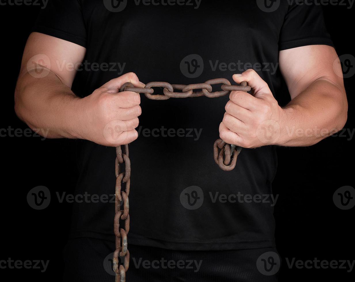 adult man in black clothes stands upright with strained muscles and holds a rusty metal chain photo