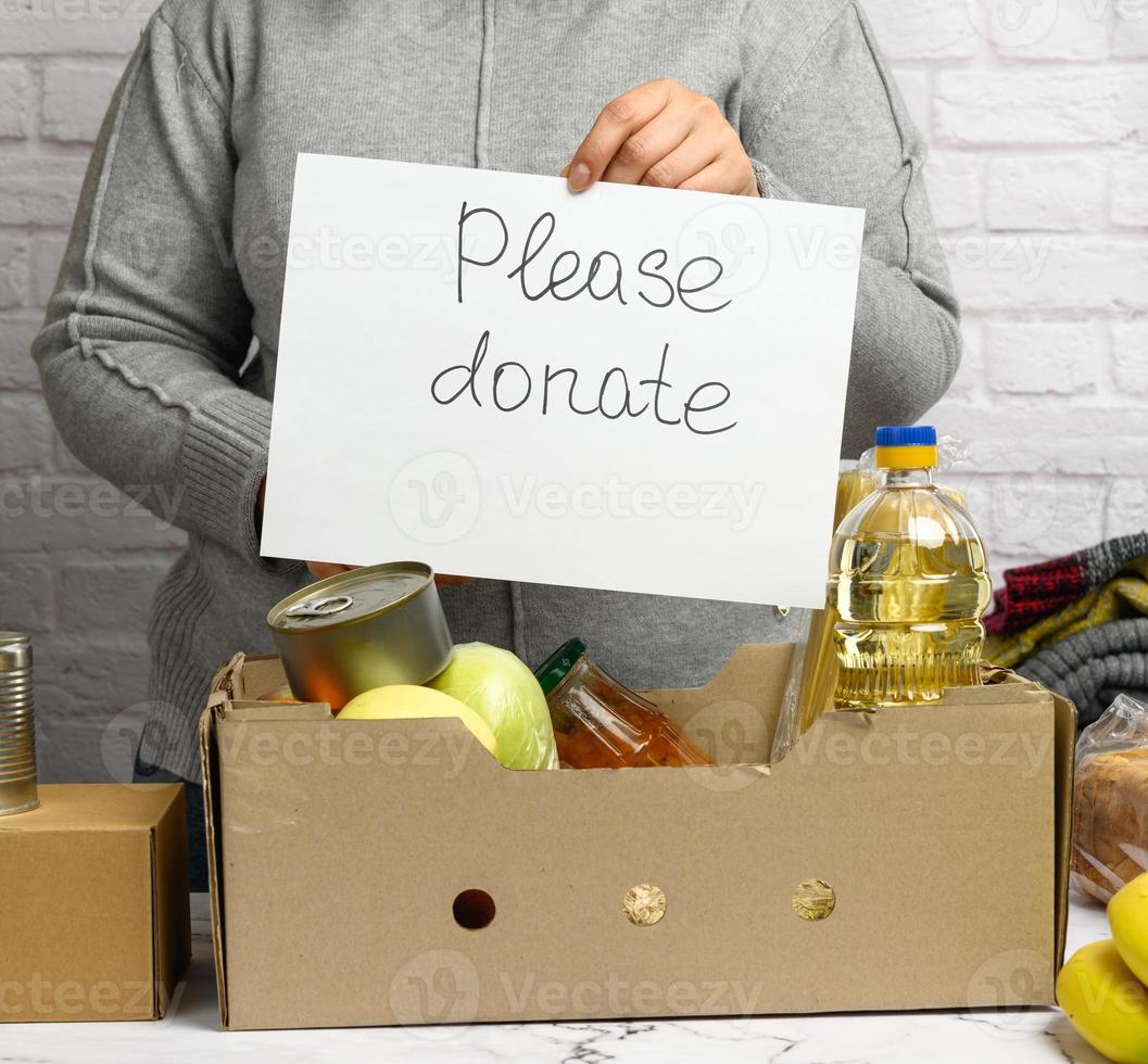 woman in a gray sweater and gloves is holding a sheet of paper with the inscription please donat, on the table there is a cardboard box with food and fruits photo