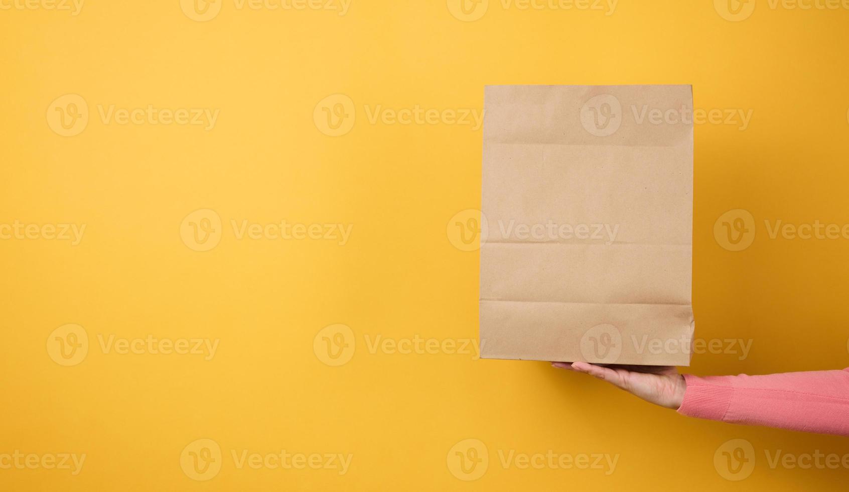 woman holding in hand brown blank craft paper bag for takeaway on yellow background. Packaging template mock up. Delivery service concept photo