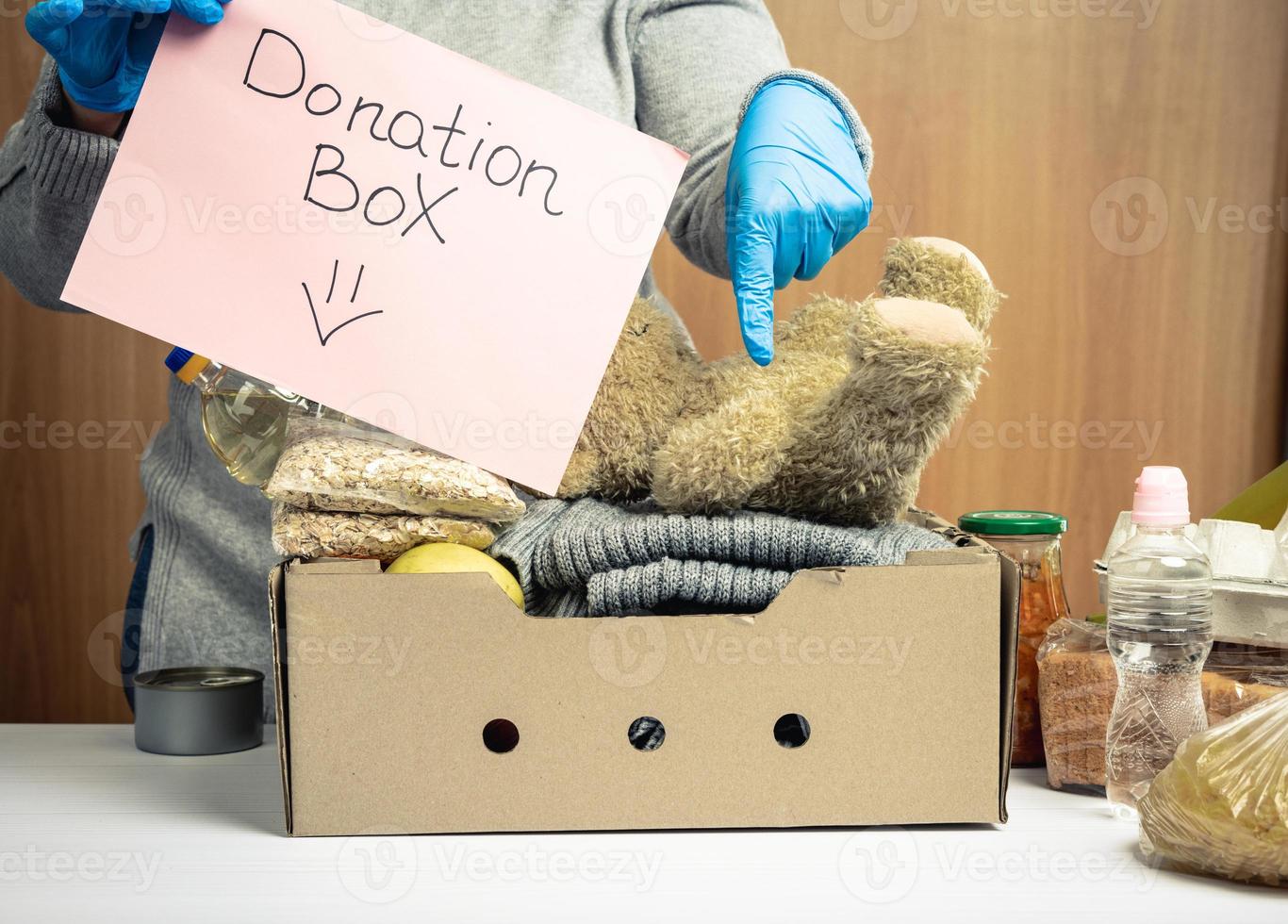 woman in gloves holds a sheet of paper with a lettering donation box and a cardboard box with food and things photo