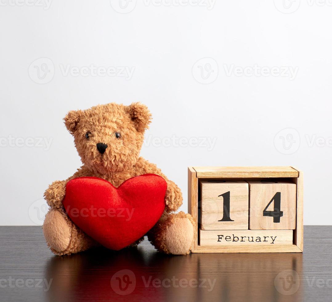 calendario de madera de cubos con la fecha del 14 de febrero y un oso de peluche marrón sobre una mesa negra foto