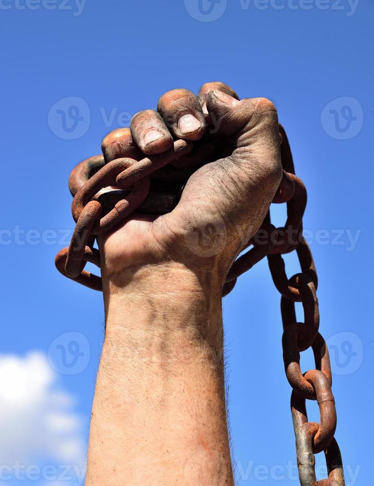 man's hand wrapped in an iron rusty chain lifted up against a blue sky photo