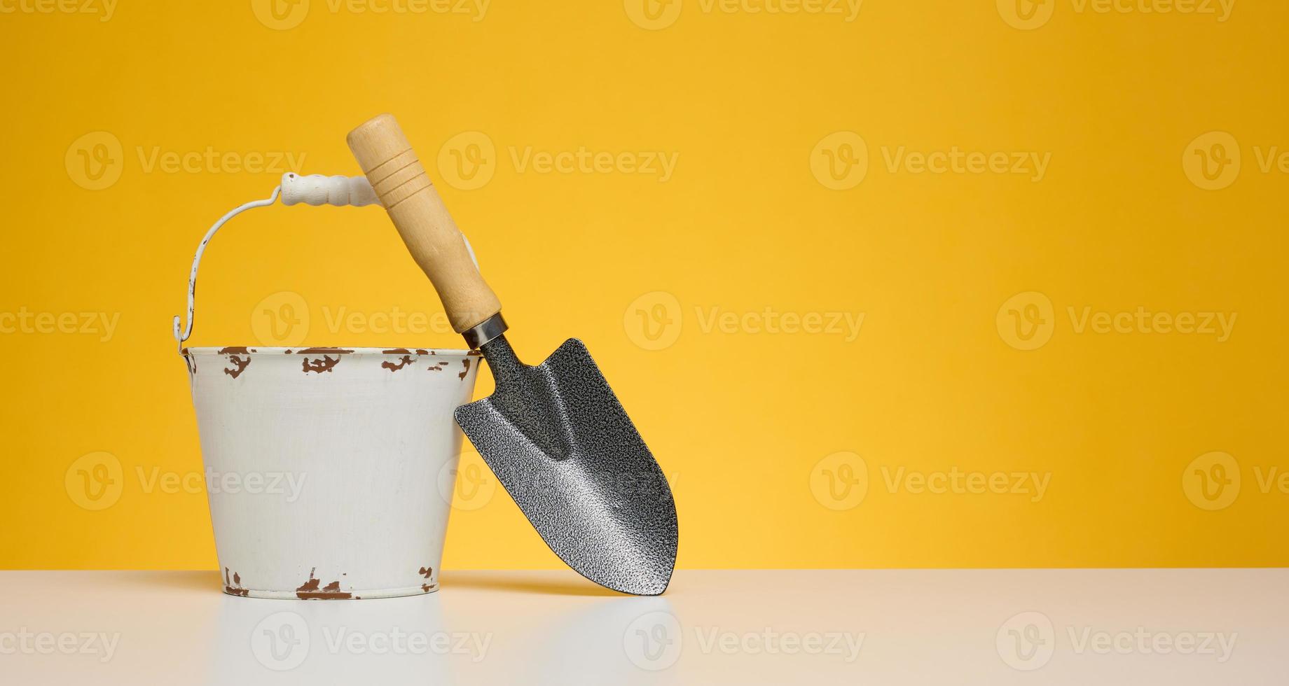 cubo de metal blanco y pala de jardín en miniatura sobre mesa blanca, fondo amarillo foto