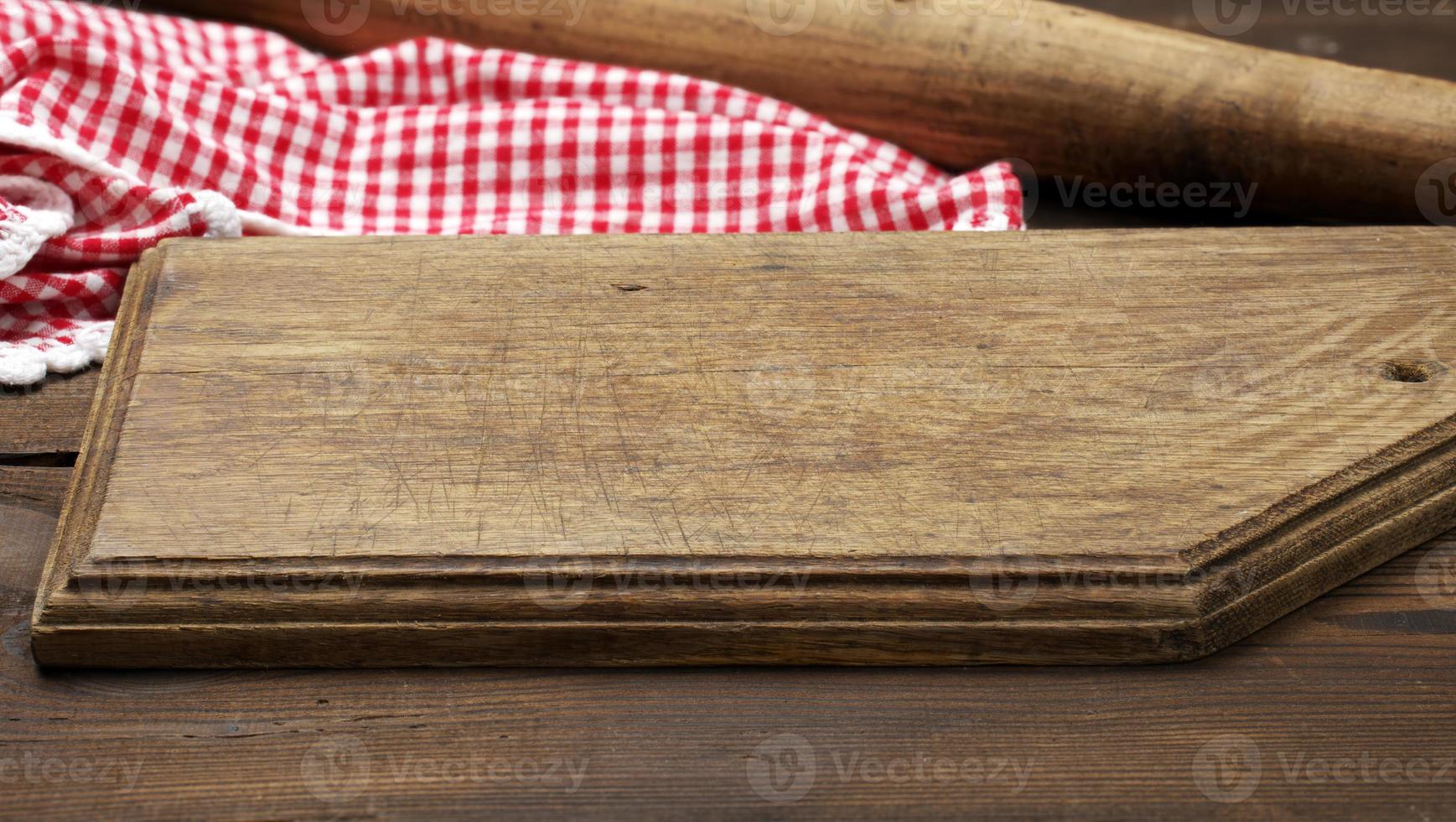 empty old wooden cutting board and folded red and white cotton kitchen napkin on a wooden brown background, top view photo