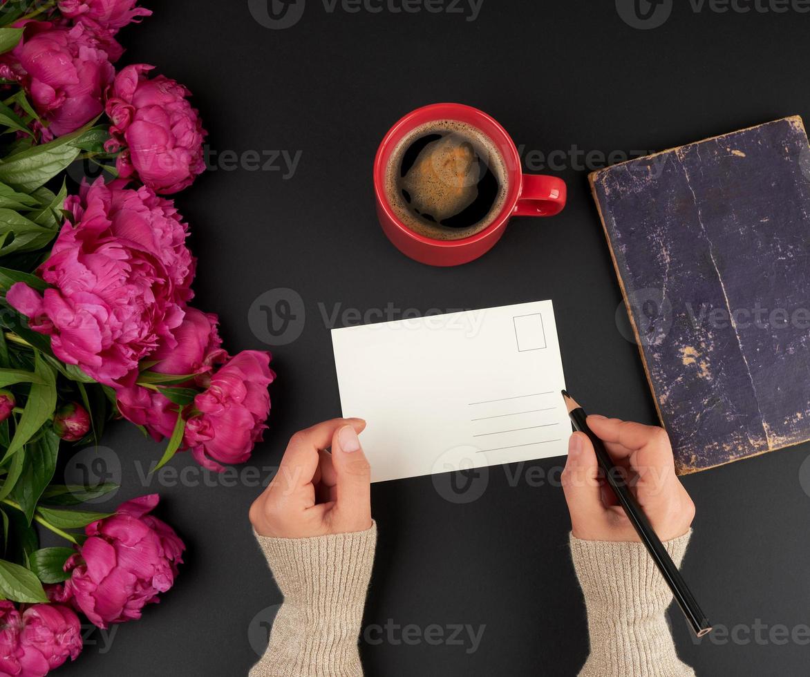 empty white postcard and two female hands, on the table a bouquet of red peonies and a ceramic cup of coffee photo