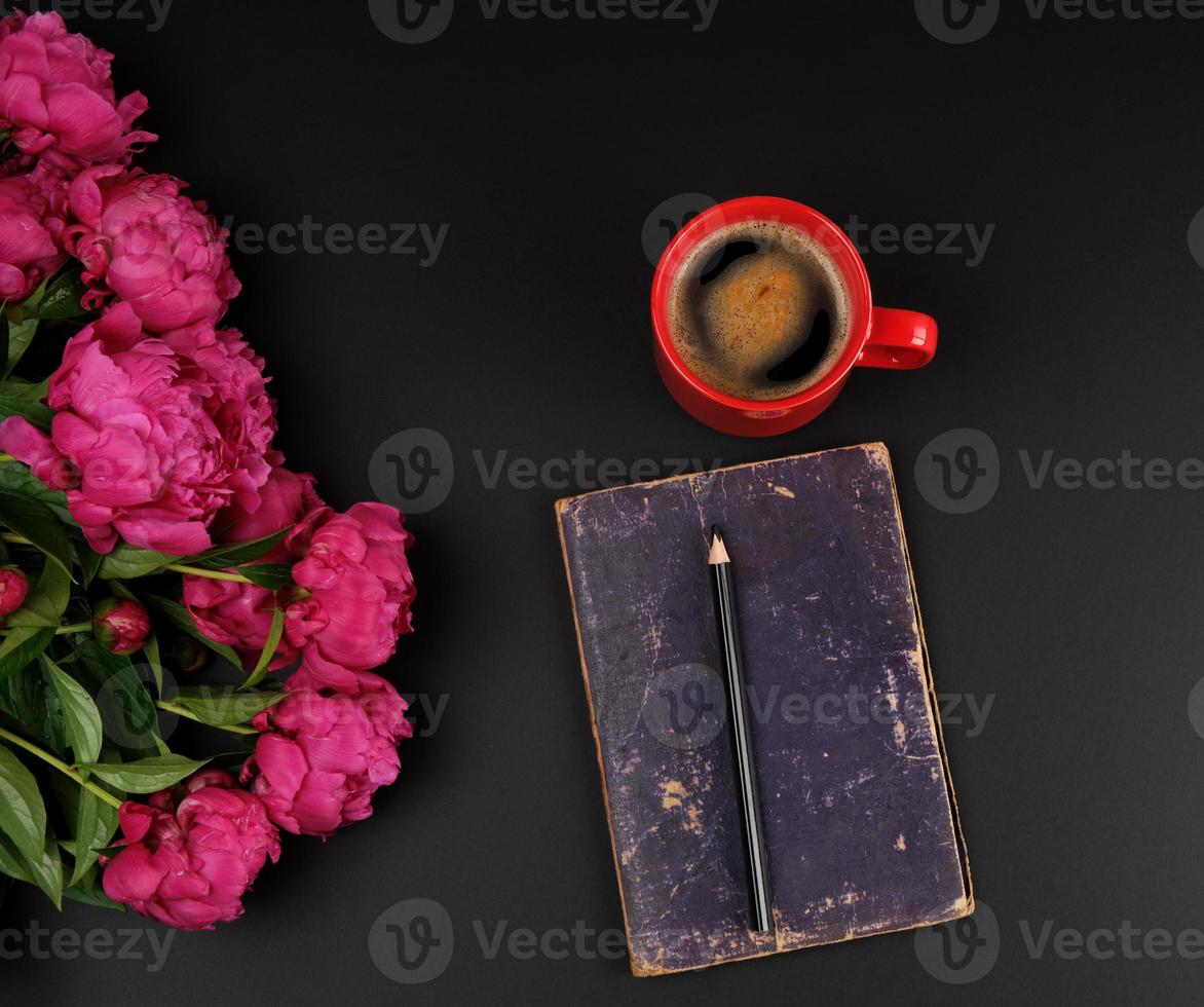 red creameric cup with black coffee, closed notebook and a bouquet of red peonies photo