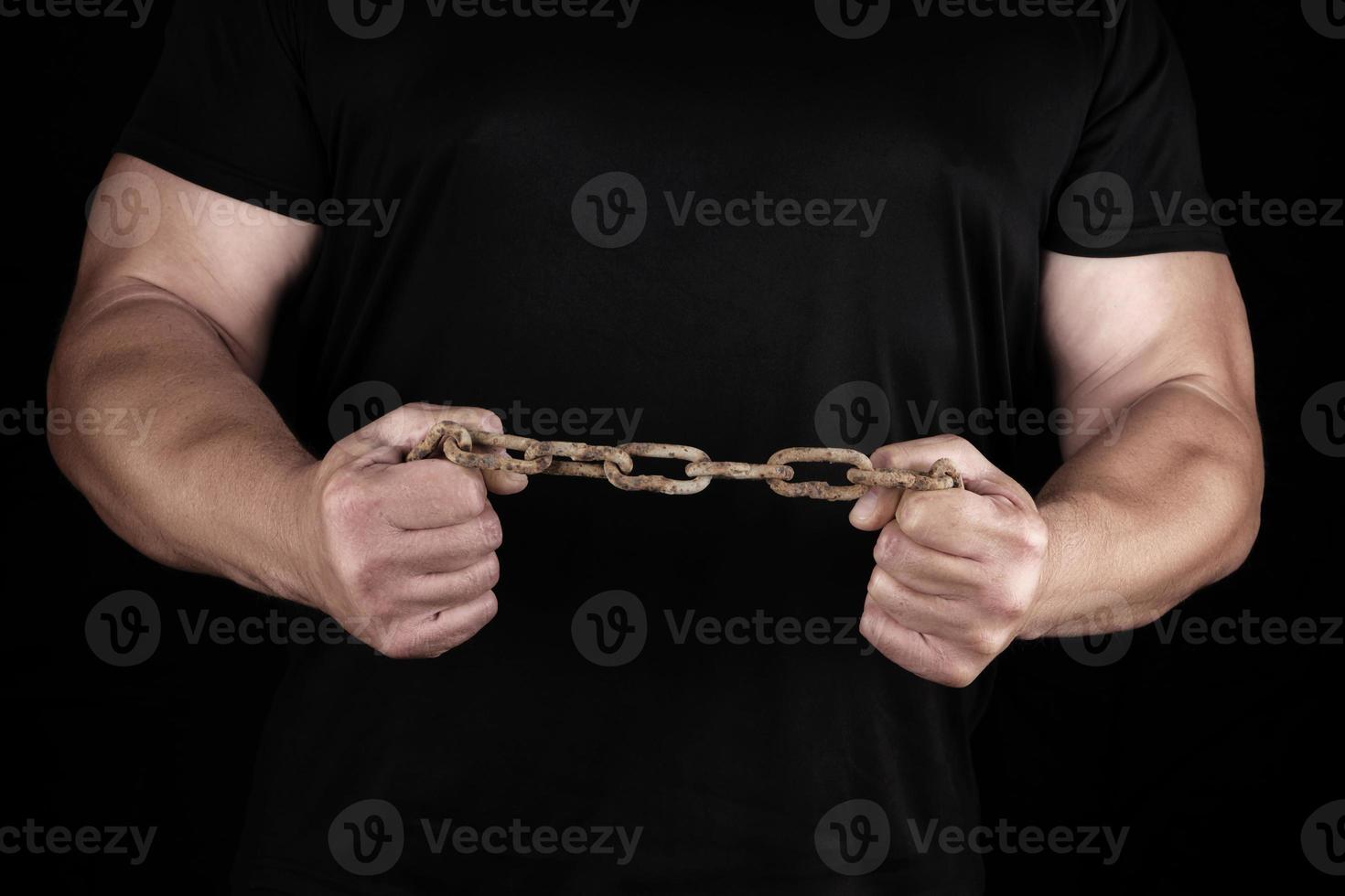 adult man in black clothes stands upright with strained muscles and holds a rusty metal chain photo