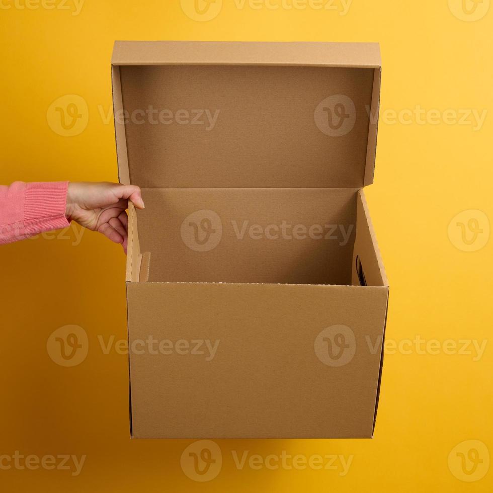 female hand holding an open brown cardboard box on a yellow background, delivery photo