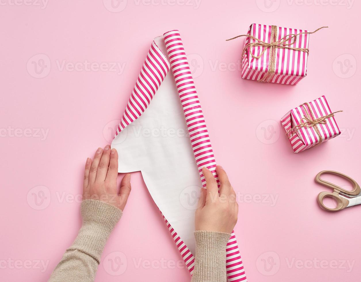 two female hands unfold a roll of wrapping paper, next to two gifts, pink background photo