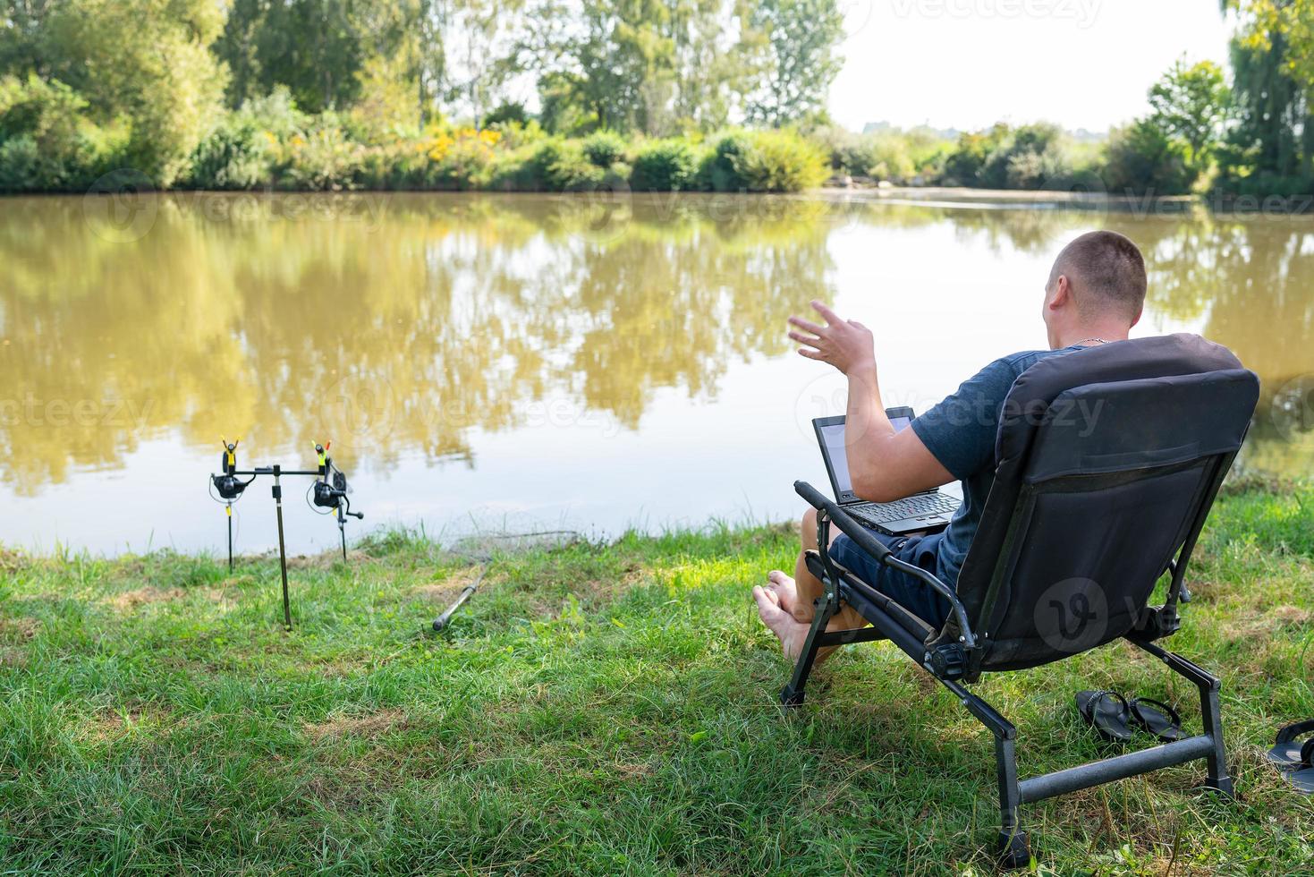 A man sits in a chair near the autumn lake and works on a laptop freelance camping. Robot out of the office, phoned with colleagues. photo