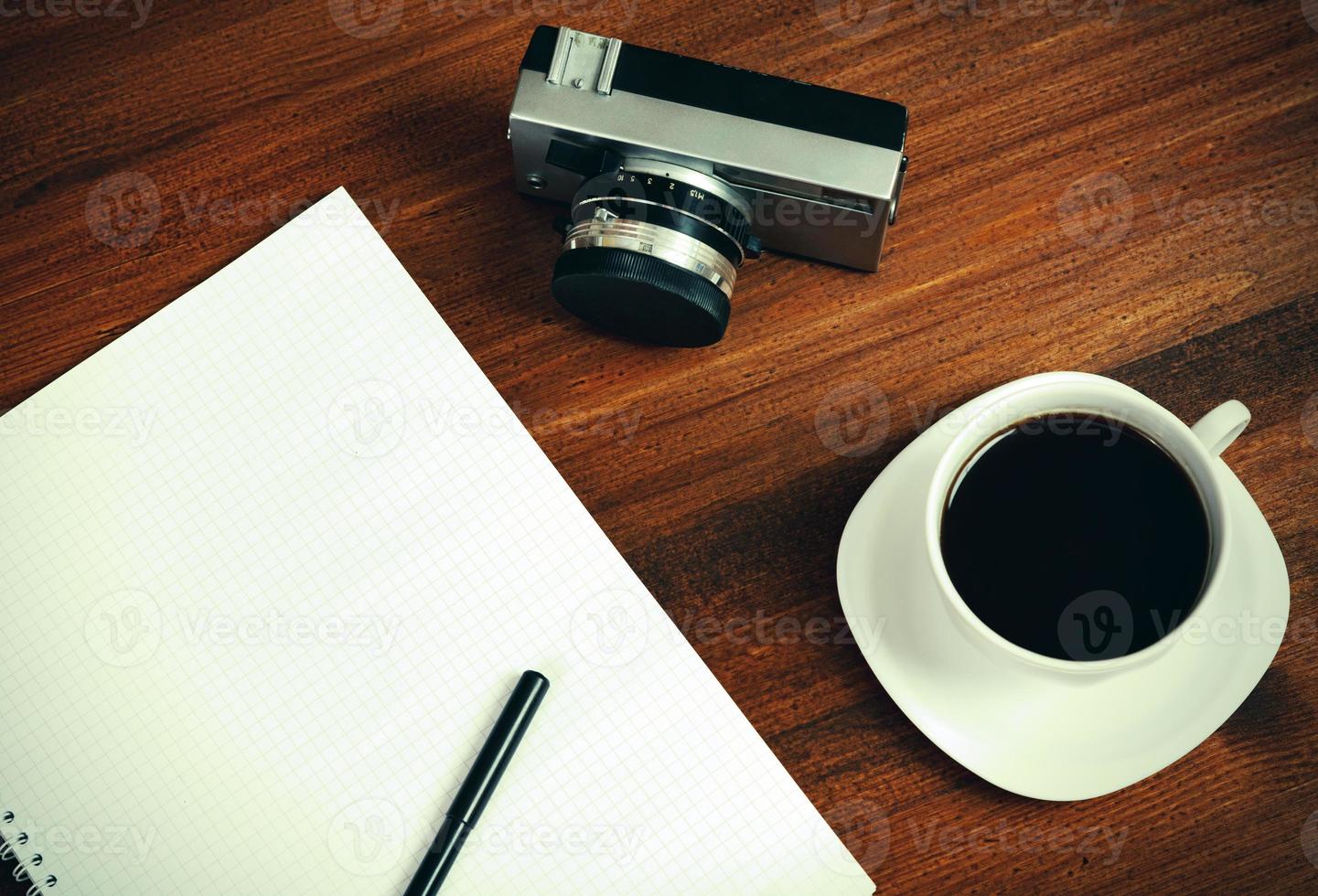 Retro film photo camera, cup of coffee and notebook with pencil on brown table. Top view.