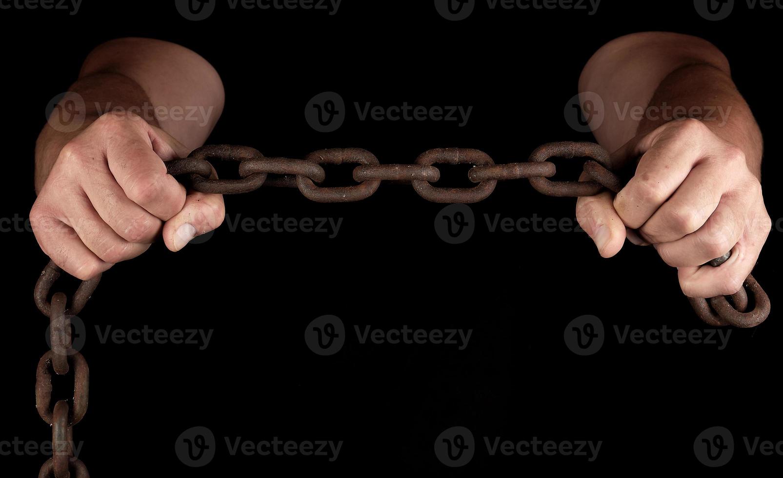 adult man in black clothes stands upright with strained muscles and holds a rusty metal chain photo