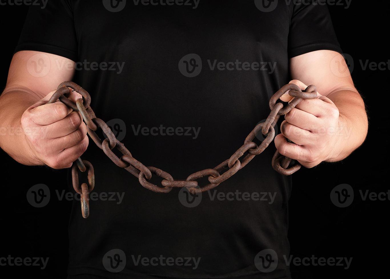 adult man in black clothes stands upright with strained muscles and holds a rusty metal chain photo