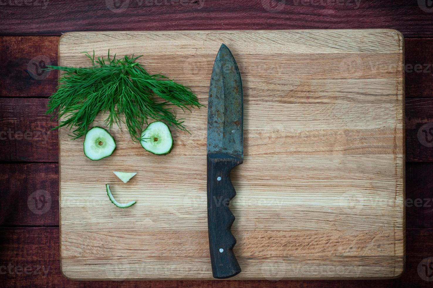 Funny faces of cucumber slices and dill on the chopping board with a knife photo