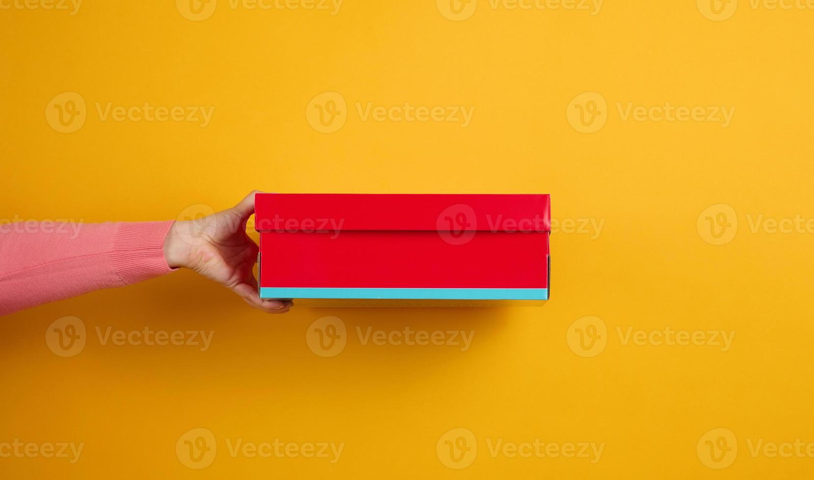 female hand holding a red cardboard box on a yellow background, delivery photo