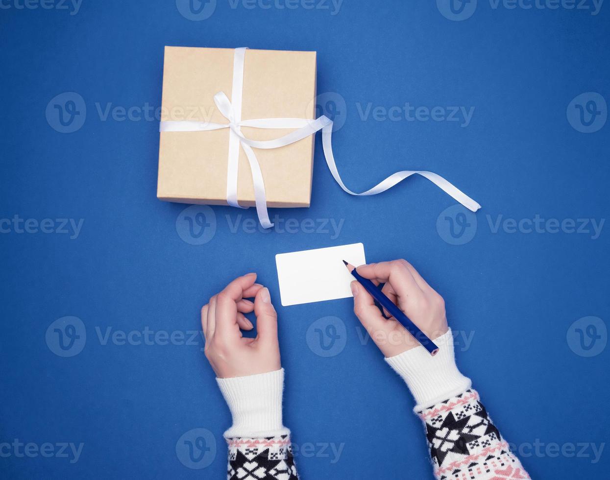 woman holds in her hand a blue wooden pencil and signs a white paper business card, next to a wrapped gift photo